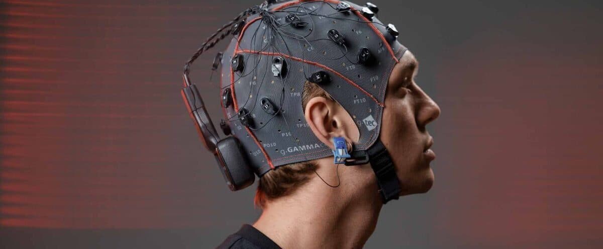 A man in profile wearing a grey EEG cap with electrodes, a wireless amplifier at the back of the cap and a blue reference clip on his ear