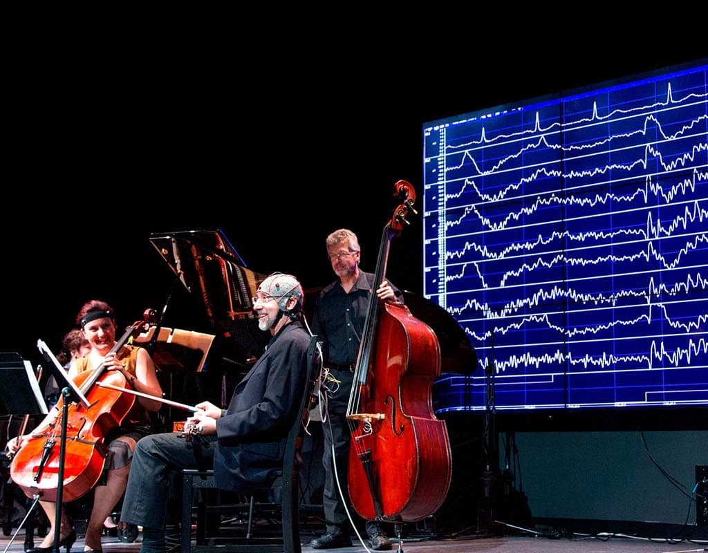 A musician wearing an EEG cap on stage beside a cellist and a double bass player, with EEG traces behind them