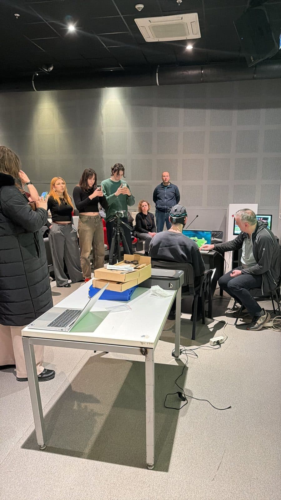 A participant wearing an EEG cap plays a BCI game on a laptop while the students around them film on their phones.