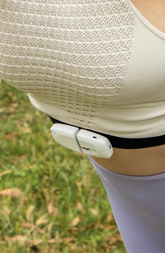 A white respiBAN respiration sensor clipped to a black band under a sports top, at chest height on someone standing outdoors